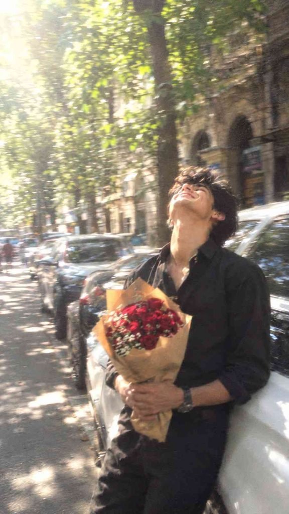 Young South Asian man holding red roses beside a white car on a sunny city street